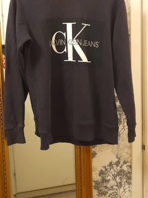 Svart sweatshirt från Calvin Klein Jeans - Svart sweatshirt med stor vit CK-logga och texten Calvin Klein Jeans på bröstet. Klassisk rund halsringning och ribbade muddar vid ärmslut och nederkant. 