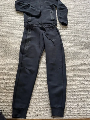 Svarta Nike Tech Fleece mjukisbyxor och hoodie  - Svarta Nike Tech Fleece mjukisbyxor med dragsko i midjan och ribbade muddar vid benslut. Byxorna har två dragkedjeförsedda fickor och diskret Nike-logga på låret. Tillverkade i mjukt och stretchigt fleecematerial för maximal komfort. Och hoodie har storlek xs medans byxorna har S 