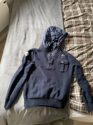 Cardigan från N.Z.A - Mörkblå cardigan med quiltad huva och knappslå framtill. Har en bröstficka med lock och snyggt broderi på bröstet samt patch på ärmen. Materialet är stickad bomull och huvan är fodrad för extra komfort. Perfekt för dig som gillar outdoor-vibe.