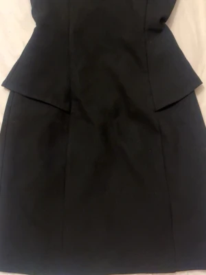 Svart figursydd peplumklänning - Snygg svart klänning med peplumdetalj i midjan och figurnära passform. Klänningen är kort och har en stilren design utan mönster. Perfekt för dig som gillar klassiska och eleganta plagg.