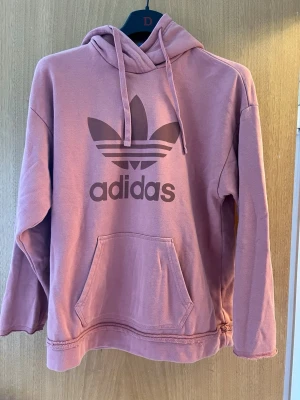 Rosa Adidas hoodie med stor logga - Säljer en rosa hoodie från Adidas med stor tryckt Trefoil-logga på bröstet. Hoodien har huva med dragsko och en stor magficka framtill. Tillverkad i mjukt bomullsmaterial och har avslappnad passform. Perfekt för en chill och sportig look.