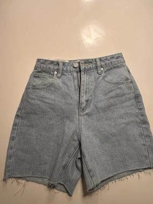 Ljusblå denimshorts med rå kant Abrand Jeans - Säljer ett par ljusblå denimshorts med hög midja och rå, fransig kant. Klassisk femficksmodell med bälteshällor och knappgylf. Perfekta för sommaren och ger en avslappnad vibe till din outfit.