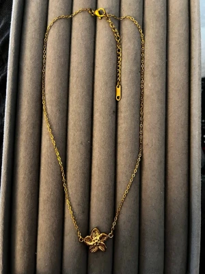 Guldigt halsband med blomma - Snyggt halsband i guldfärgad metall med en detaljrik blomma som hänge. Kedjan är tunn och har justerbar längd med karbinlås. Perfekt för dig som gillar stilrena och feminina accessoarer med en touch av naturinspiration.