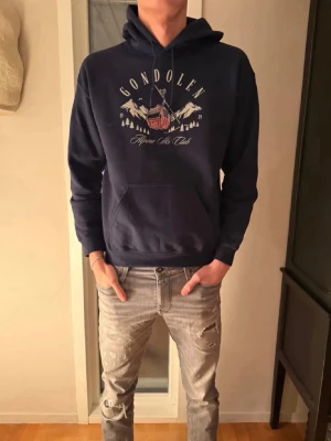 Mörkblå hoodie Gondolen Alpine Ski Club - Mörkblå hoodie med tryck av en gondol och berg samt texten 'Gondolen Alpine Ski Club' på bröstet. Klassisk känguruficka framtill och justerbar huva. Tillverkad i mjukt bomullsmaterial, perfekt för chill dagar.
