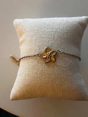 Guldigt armband med blomma 18k guldplaterat - Snyggt armband i guldfärgad 18k planterad metall med en detaljrik blomma i mitten. Kedjan är tunn och har justerbar längd.  Perfekt för dig som gillar stilrena och feminina accessoarer.