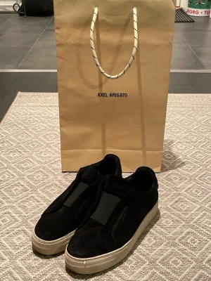 Svarta sneakers från Axel Arigato - Stilrena svarta sneakers från Axel Arigato i mocka med vit sula och slip-on design utan snörning. Skorna har en minimalistisk look och bred elastisk panel upptill för enkel påtagning. Perfekta för dig som gillar en clean och modern stil. Dustbag fås med.