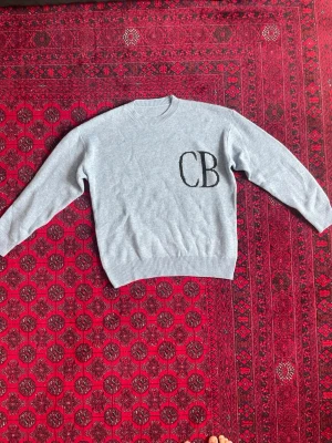 Blå knitted sweater  - Säljer en ljusgrå stickad tröja med rund hals och ribbade muddar. På bröstet finns en stor svart CB-logga som ger en cool och personlig touch. Perfekt för dig som gillar enkel men snygg stil.