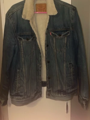 Levi's fodrad jeansjacka blå M - Säljer en klassisk blå jeansjacka från Levi's med vit teddyfoder på insidan och kragen. Jackan har två bröstfickor med lock, knäppning framtill och raka ärmar. Perfekt för dig som gillar en tidlös och cool stil.