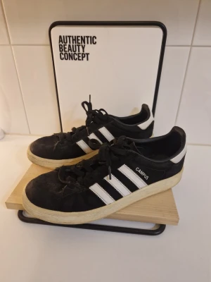 Svarta Adidas Campus sneakers - Klassiska Adidas Campus sneakers i svart mocka med vita detaljer och de ikoniska tre ränderna på sidan. Låg modell med vit sula och snörning. Perfekta för en avslappnad streetwear-look.
