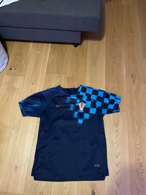 Mörkblå Kroatien fotbollströja Nike - Snygg mörkblå fotbollströja från Nike med Kroatiens landslagsemblem på bröstet och röd Nike-logga. Tröjan har ett rutigt mönster i ljusblått över axeln och är tillverkad i ett lätt, snabbtorkande material. Perfekt för träning eller match.