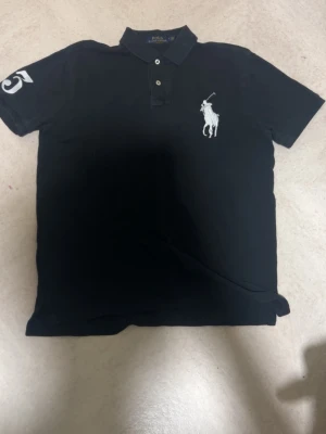 Svart pikétröja från Polo Ralph Lauren - Svart pikétröja från Polo Ralph Lauren med klassisk krage, knäppning med tre knappar och stor vit broderad logga på bröstet. Siffra 3 på ärmen. Tillverkad i mjuk bomull och passar perfekt till en avslappnad stil.