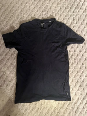 Mörkblå t-shirt från Jack & Jones - Snygg mörkblå t-shirt från Jack & Jones Essentials i ekologisk bomull. Klassisk modell med rund halsringning och en liten bröstficka. Perfekt basic-plagg att ha i garderoben. Hör av dig vid frågor!
