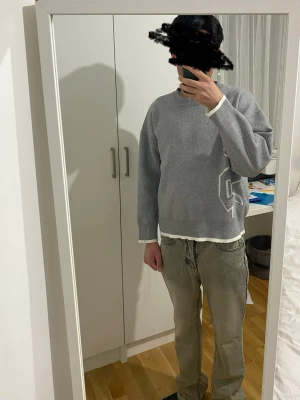 Grå stickad tröja med vit detalj - Snygg grå stickad tröja med en stor vit grafisk detalj på sidan. Tröjan har rund hals, långa ärmar och ribbade muddar vid ärmslut och nederkant. Perfekt för lager-på-lager och enkel att matcha med jeans.