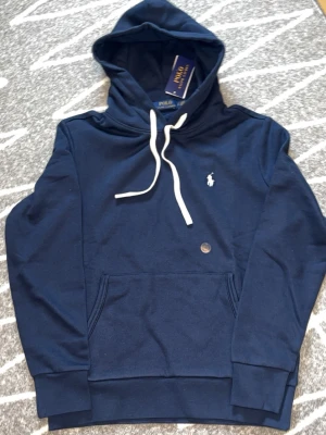 Mörkblå hoodie från Polo Ralph Lauren - Mörkblå hoodie från Polo Ralph Lauren med vit dragsko och klassisk broderad logga på bröstet. Hoodien har känguruficka framtill och ribbade muddar vid ärmslut och nederkant. Perfekt för en chill och stilren look.