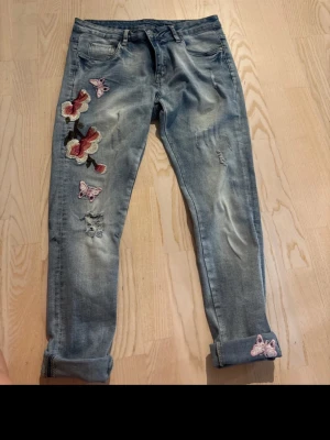 Blå jeans med blommor och fjärilar - Snygga ljusblå jeans med broderade blommor och fjärilar på vänster ben. Jeansen har en avslappnad passform, uppvikta benslut och slitningar för en trendig look. Perfekta för dig som vill sticka ut lite extra. Femficksmodell och klassisk dragkedja. Men vet ej vart de är ifrån 