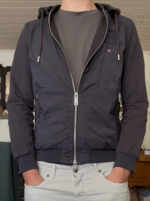 Hybridjacka Aeronautica Militare - Snygg blå vindjacka från Aeronautica Militare med avtagbar huva och dubbelzip framtill. Jackan har ribbade muddar, två fickor med dragkedja. Schyssta armbågspatches och huvsnören. Italiensk strl - S mer som XS.