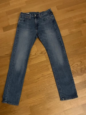 Levi's raka blå jeans - Klassiska blå jeans från Levi's med rak passform och fem fickor. Jeansen har en snygg tvätt med lätt slitning på låren och bakficka med den ikoniska röda Levi's-taggen. Perfekta för en avslappnad och tidlös stil.