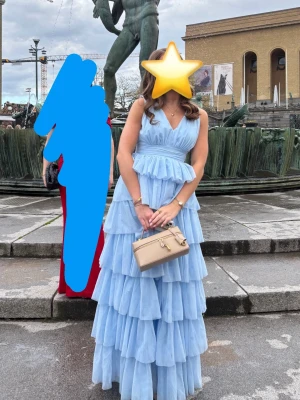 Ljusblå volangklänning med v-ringning - Säljer en lång, ljusblå klänning med flera lager volanger i tyll och v-ringad hals. Klänningen har markerad midja och är ärmlös. Från Bubbleroom , nypris 1800kr. Perfekt för tex bal eller bröllop. Använd två gånger och är i mycket bra skick!