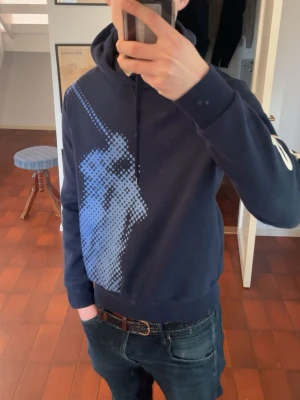 Mörkblå ralph lauren hoodie - Snygg mörkblå hoodie från Ralph Lauren med stort pixelmönstrat tryck av en polospelare på framsidan och vit text längs ena ärmen. Klassisk huva med dragsko och ribbade muddar. Perfekt för dig som gillar streetwear och sportig stil. Jättebra skick.