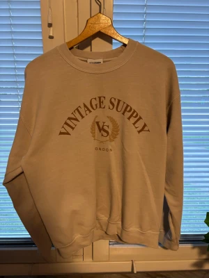 Beige sweatshirt från Vintage Supply - Beige sweatshirt från Vintage Supply med trycket 'VINTAGE SUPPLY VS LONDON' i brunt på bröstet. Tröjan har rund halsringning, långa ärmar och ribbade muddar vid ärmslut och nederkant. Tillverkad i mjukt bomullsmaterial för en avslappnad look.