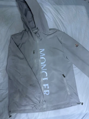  Moncler vindjacka  - Snygg ljusgrå vindjacka från Moncler med stor logga framtill och klassisk Moncler-patch på ärmen. Jackan har huva, dragkedjor på fickorna och justerbar nederkant. Materialet är tunt och smidigt, perfekt för blåsiga dagar. Helt ny aldrig använt 