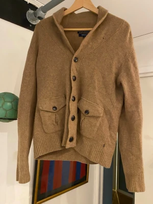 Beige kofta i merinoull från Gant - Snygg beige kofta från Gant i extrafin merinoull. Koftan har stora knappar framtill, två fickor med lock och ribbade muddar vid ärmslut och nederkant. Perfekt för lager-på-lager och en chill stil.