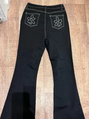 Svarta utsvängda jeans med kontrastsömmar - Svarta jeans med utsvängda ben och vita kontrastsömmar. Bakfickorna har broderade blommor som ger en cool retrovibe. Klassisk femficksmodell med knapp och dragkedja framtill. Perfekta för dig som gillar 70-talsinspirerad stil.