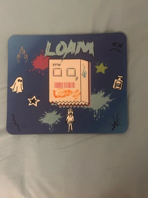 LOAM musmatta med grafiskt tryck - Cool musmatta med blå bakgrund och grafiskt tryck med texten LOAM, tecknade figurer och färgstänk. Rektangulär form med rundade hörn, ovansida i tyg och undersida i svart gummi för bra grepp. Perfekt för gaming eller datorarbete.