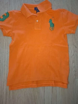 Orange polotopp från Ralph Lauren - Cool orange polotopp från Ralph Lauren med klassisk krage och två knappar. Stort grönt broderat logomärke på bröstet och en grön siffra på ärmen. Perfekt för dig som gillar sportig och stilren look. Den är stol s men kan va lite mindre