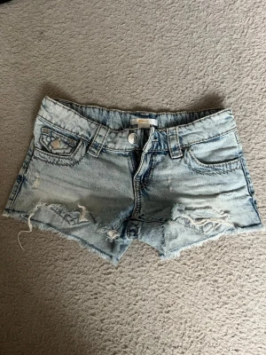Ljusblåa jeansshorts - Snygga ljusblåa jeansshorts från Ginatricot med slitna detaljer och råa kanter. Shortsen har klassiska fem fickor, broderade bakfickor och silverfärgade knappar. Perfekta för en avslappnad och trendig look.