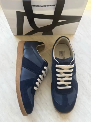 Masion margiela gats  - Snygga mörkblå sneakers från Maison Margiela med ovandel i läder och mocka, vita snören och klassisk gummisula i brunt. Modellen har en retroinspirerad design med låg profil och diskreta paneler i olika material. Perfekta för dig som gillar stilrena och tidlösa sneakers.