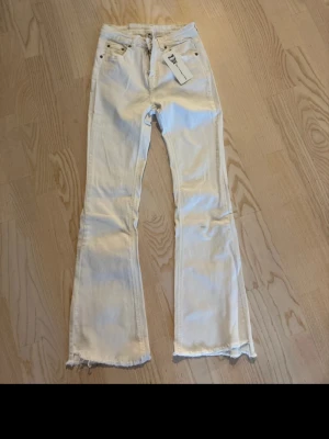 Vita bootcut jeans från  - Säljer ett par vita bootcut jeans med råa kanter nertill och klassisk femficksdesign. Jeansen har hög midja och är tillverkade i ett mjukt denimtyg. Perfekta för dig som vill ha en clean och trendig look.