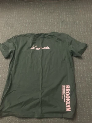 Grön t-shirt med Brooklyn-tryck - Mörkgrön t-shirt från Keymate med vit text på bröstet och rosa Brooklyn-tryck nedtill. Klassisk passform och rund halsringning. Perfekt för dig som gillar streetwear och vill ha en enkel men snygg look. Storlek 164
