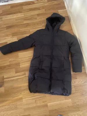 Svart lång dunjacka med huva - Säljer en svart lång dunjacka från Peak Performance med huva och dragkedja framtill. Jackan har två fickor med dragkedja och är perfekt för kalla vinterdagar. Klassisk puffer-modell som håller dig varm och stilren.