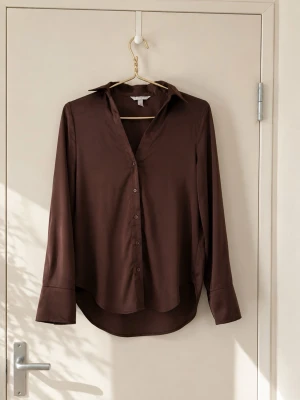 Brun skjorta med långa ärmar - Stilren brun skjorta med klassisk krage och långa ärmar. Skjortan har knappar framtill och breda manschetter. Tillverkad i ett mjukt, slätt material som ger en snygg och enkel look.Finns extra 2 knappar. 