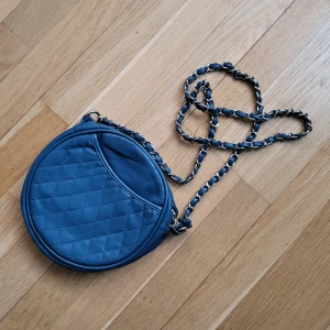 Liten blå rund handväska - Inköpt mellan år 2000-2010. Liten och blå handväska med guldig kedja längs axelremmen. Jättefint skick utanpå, nyskick inuti. Kan ta fler bilder och kolla detta. Svarar på alla frågor.