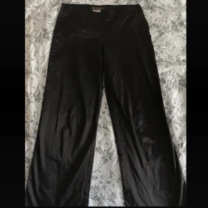 Lågmidjade raka kostymbyxor  - Vida/ raka kostymbyxor med glansigt svart tyg och låg midja, på ena sidan så har byxan gått lite sönder, men det går att sy🤍 Mått:  ⚜️Midja 76 cm ⚜️Längd: 100 cm