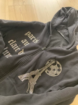 Svart luvtröja Blinglucid - Oanvänd ny svart luvtröja med zip. Rhinestone i mönster. Storlek XS men mer lik en M. Inköpt för 699 kr.