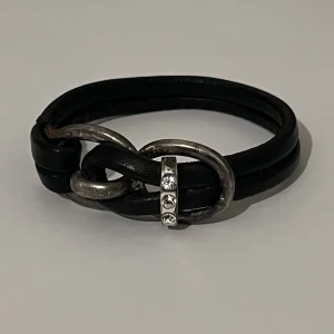 Svart armband  - Svart armband i läder (eller fakeläder) och ett spänne i metall med stenar.