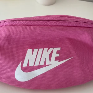 Midjeväska Nike - Midjeväska från Nike. Finns tecken på användning i form av några grå streck i tyget (se bild tre för exempel). Väskan är rymlig och har ett huvudfack med ett mindre fack, sam ett fack på baksidan