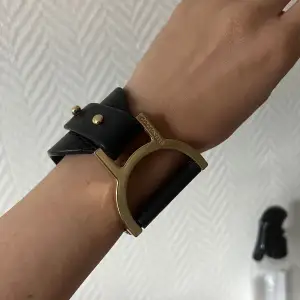 Svart armband från Coccinelle i modell ”Arlettis” i skinn med guldfärgade detaljer.  Ordinarie pris 700:-