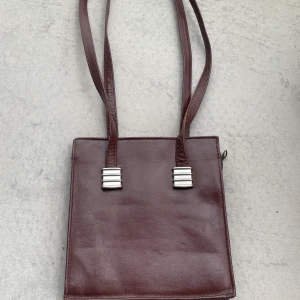 Väska - Rödbrun, fyrkantig väska med silverdetaljer, i läder. Två straps, som sitter bekvämt över ena axeln. I bra skick! Passar bra om man ska ut och vill ha en liten smidig väska, kan skicka mått på förfrågan!