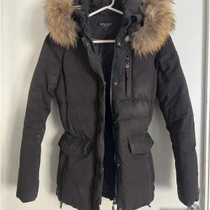 Hollies Jacka - Storlek 34 . Dragtråden för att justera midjan på ena sidan är trasig men det går bra att justera med andra sidan. Väldigt varm och praktisk jacka med äkta päls vid luvan. 6 fickor totalt ( två vid nederdelen , en vid bröstet och  en på insidan. 