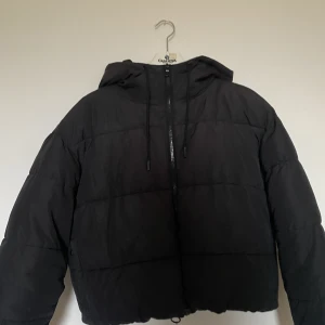 Croppad vinterjacka - Svart croppad vinterjacka från Monki i storlek S. Jackan har luva och resårband längst ner som man kan dra i för att ändra i midjan. Jackan har två fickor i sidorna och en innerficka. Använd mycket men inga tecken på slitage. 