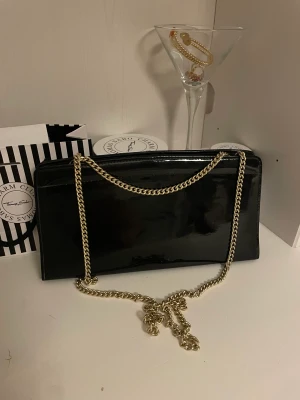 SVART GLANSIG AXELREMSVÄSKA MED GULDIG KEDJA - Kuvertväska, liten väska med guldig kedja. Perfekt storlek till fester eller middagar. Får plats med mobil, nycklar, kort och läppglans m.m. Den är i väldigt bra skick och har knappt använts. Jag köpte den för cirka 600kr. Skriv för fler frågor eller bild