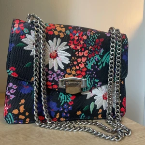Blommig väska - Blommig väska från mango med silverstraps, går att ha korta och långa.  Spännet är slitet. I övrigt bra skick. Ca 2 dm hög, 2,5 bred. Får plats med pocketbok.