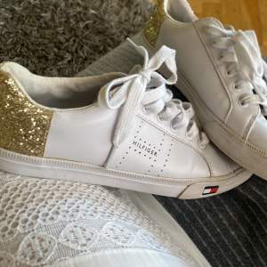 Vita sneakers från Tommy Hilfiger. Jättebra skick. I skorna står det storlek 8 1/2 vilket blir ca. 39-40 i Svensk storlek. Rekommenderar till en storlek 40. Äkta läder och har guldiga glitter på baksidan vilket passar jättebra nu till sommaren. 