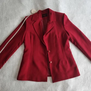Vintage elegance alert : red coat - En vintage röd kappa som gör en djärv comeback! Prydd ned charmiga röda knappar och med en vackert kammat midjeparti är denna kappa själv epitomen av retrosofistikering. Det är en del av dräkten med väst och byxor men kan också köppas separat 