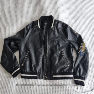 Vintage edge: svart läder sportjacka med broderi - Släpp lös din vilda sida med denna vintage svarta läder sportjacka med utsökt broderi på båda ärmarna! Omfamna den perfekta blandningen av edgy vilket gör ett djärvt uttalande var du än går.