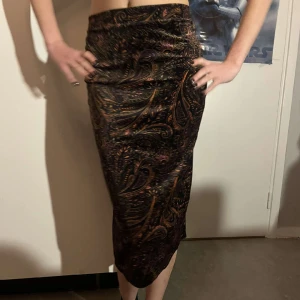 Boho midikjol mönstrad - Stretchigt material speciellt i midjan, sammet. Fint mörkt mönstrade med svart bakgrund och orange krumelurer samt lite lila och blått. Lätt använd, inget slitage.